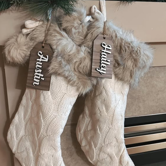 Two beige knitted stockings with fur trim and personalized tags hanging on a fireplace.