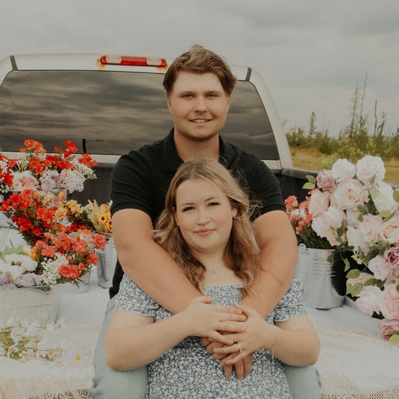 A couple posing in the bed of a pickup truck surrounded by large flower arrangements. The man wears a black polo shirt and has his arms around the woman, who is wearing a light blue and white floral dress.