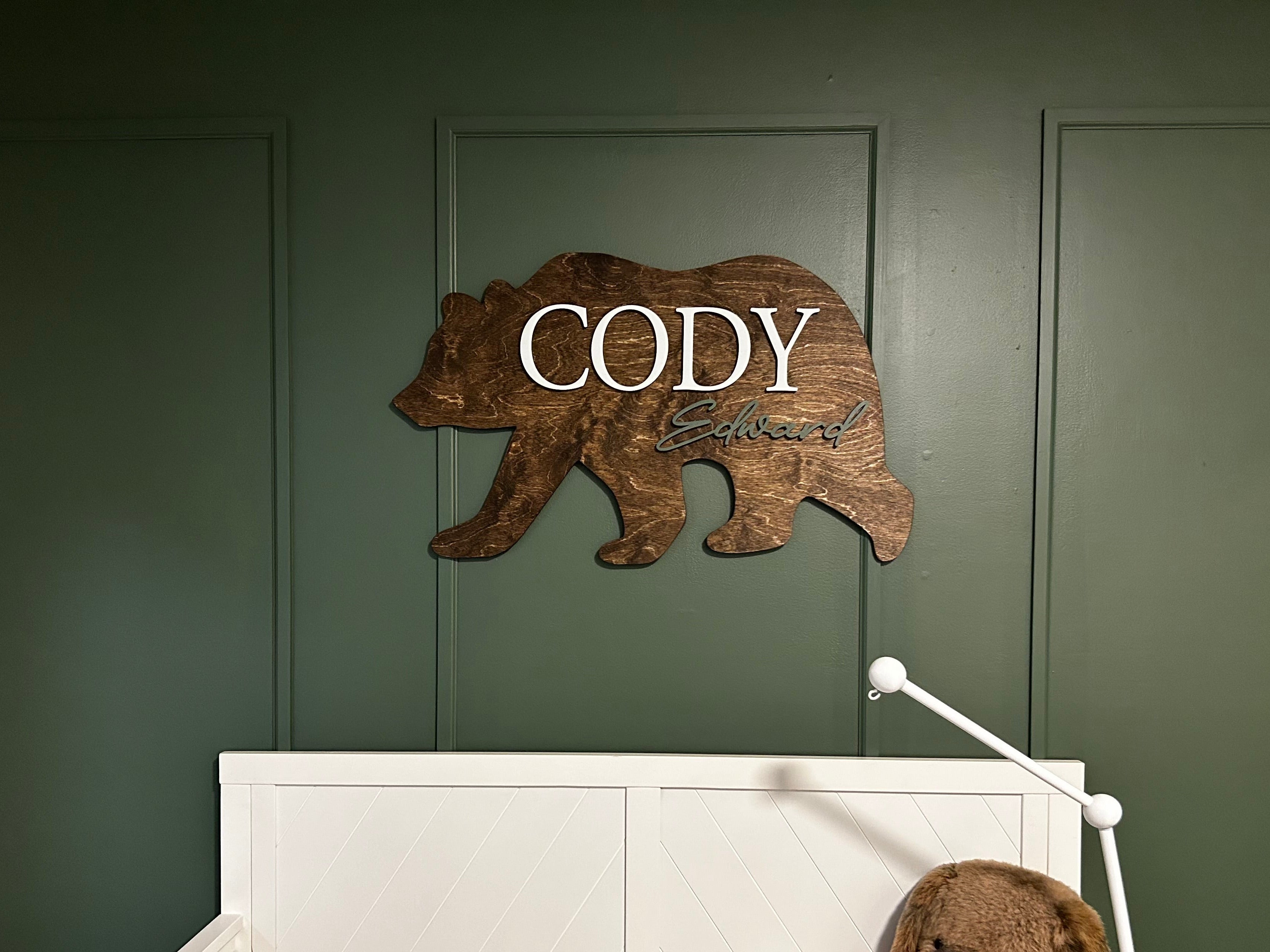 Wooden bear-shaped sign with 'CODY' on a green wall above a crib with teddy bears.