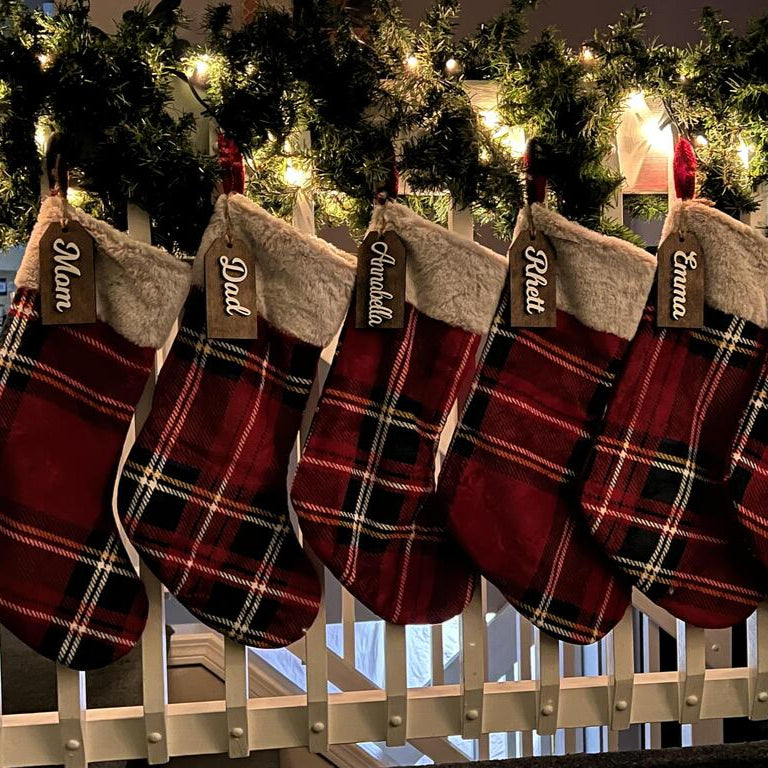Plaid Christmas stockings hanging on a white picket fence with lights and greenery in the background.